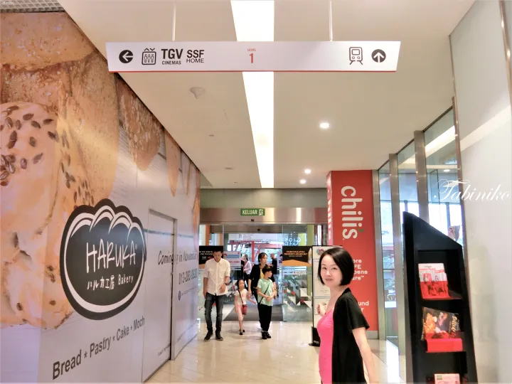TGVシネマズの案内表示の下、Haruka Bakeryの仮囲いとChili’sの看板があるショッピングモールの廊下でポーズをとる女性。