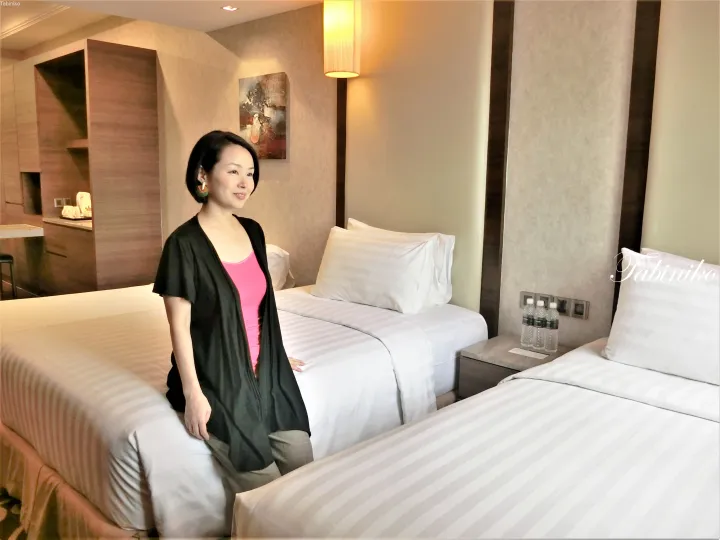 モダンなホテルの客室で、ピンクのトップスと黒のカーディガンを着た女性がベッドに座っている。