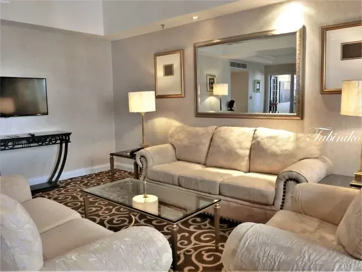 Elegant hotel suite living room with beige sofa, glass coffee table, and large mirror reflecting the interior space.