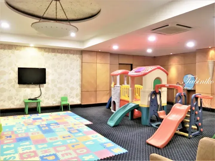 Colorful indoor children&rsquo;s play area with a plastic house, slide, alphabet mat, and TV in a hotel room.
