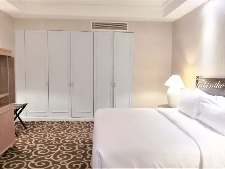 Spacious hotel bedroom with white wardrobe, patterned carpet, and neatly made bed.