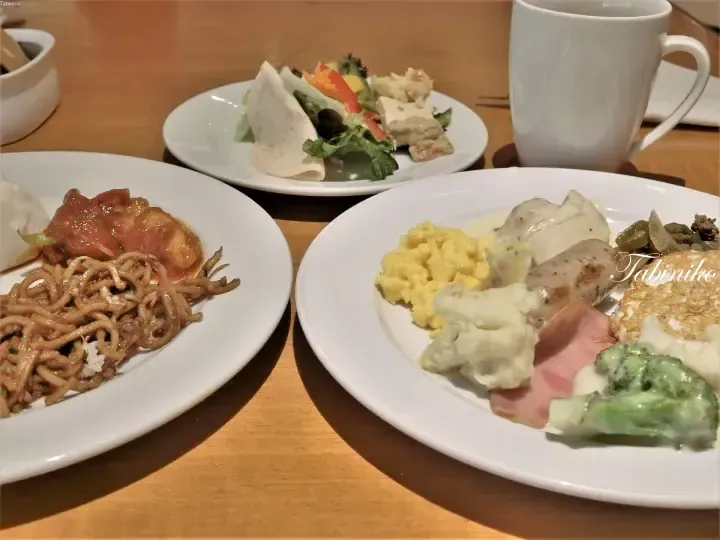 A buffet spread featuring stir-fried noodles, scrambled eggs, sausages, and a fresh salad on white plates at a dining table.
