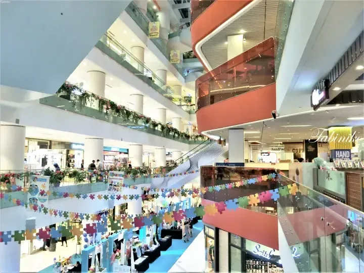 Multi-level shopping mall atrium with glass railings, escalators, and colorful puzzle piece decorations hanging across the open space.