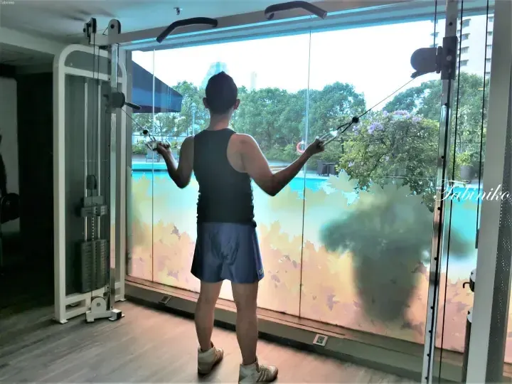 Man exercising on a cable machine at a gym with a view of a swimming pool and lush greenery outside.