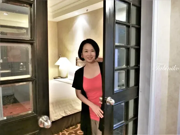 Smiling woman in pink top and black cardigan stands in doorway of a hotel room with a bed and lamp visible inside.