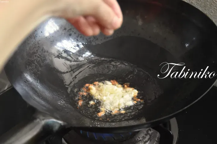 一隻手在藍色火焰的炒鍋上方撒調味料,鍋中正在翻炒蒜末和蔥頭。
