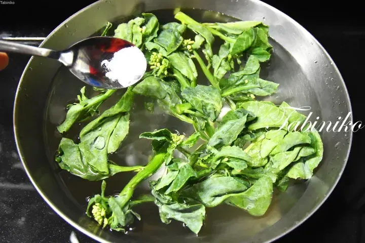 Fresh green vegetables boiling in a pot with a spoonful of salt being added.