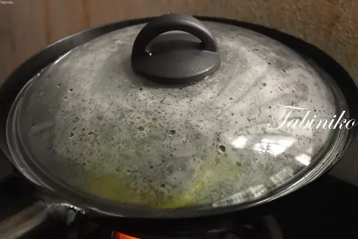 A close-up of a glass pot lid covered in condensation, revealing green vegetables simmering inside on a stove.